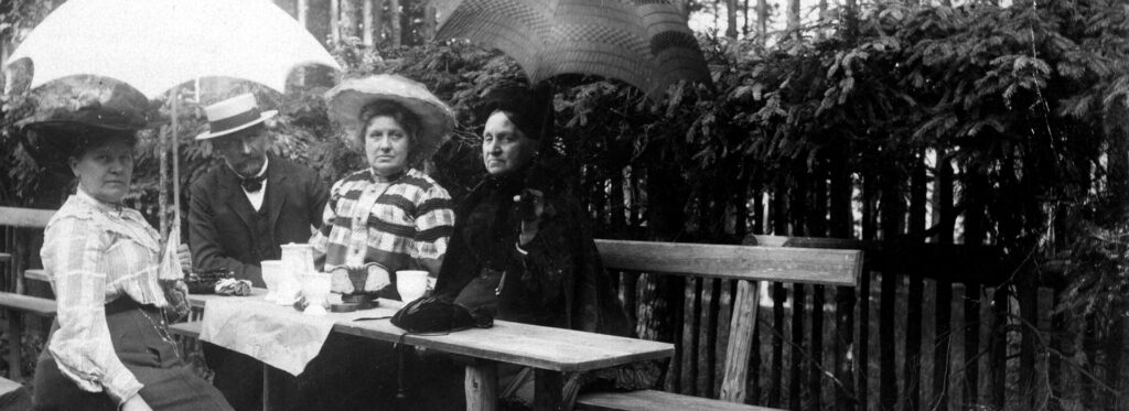 Gruppe von drei Damen und einem Herren auf einer Holzbank bei Kaffee und Kuchen, um 1900